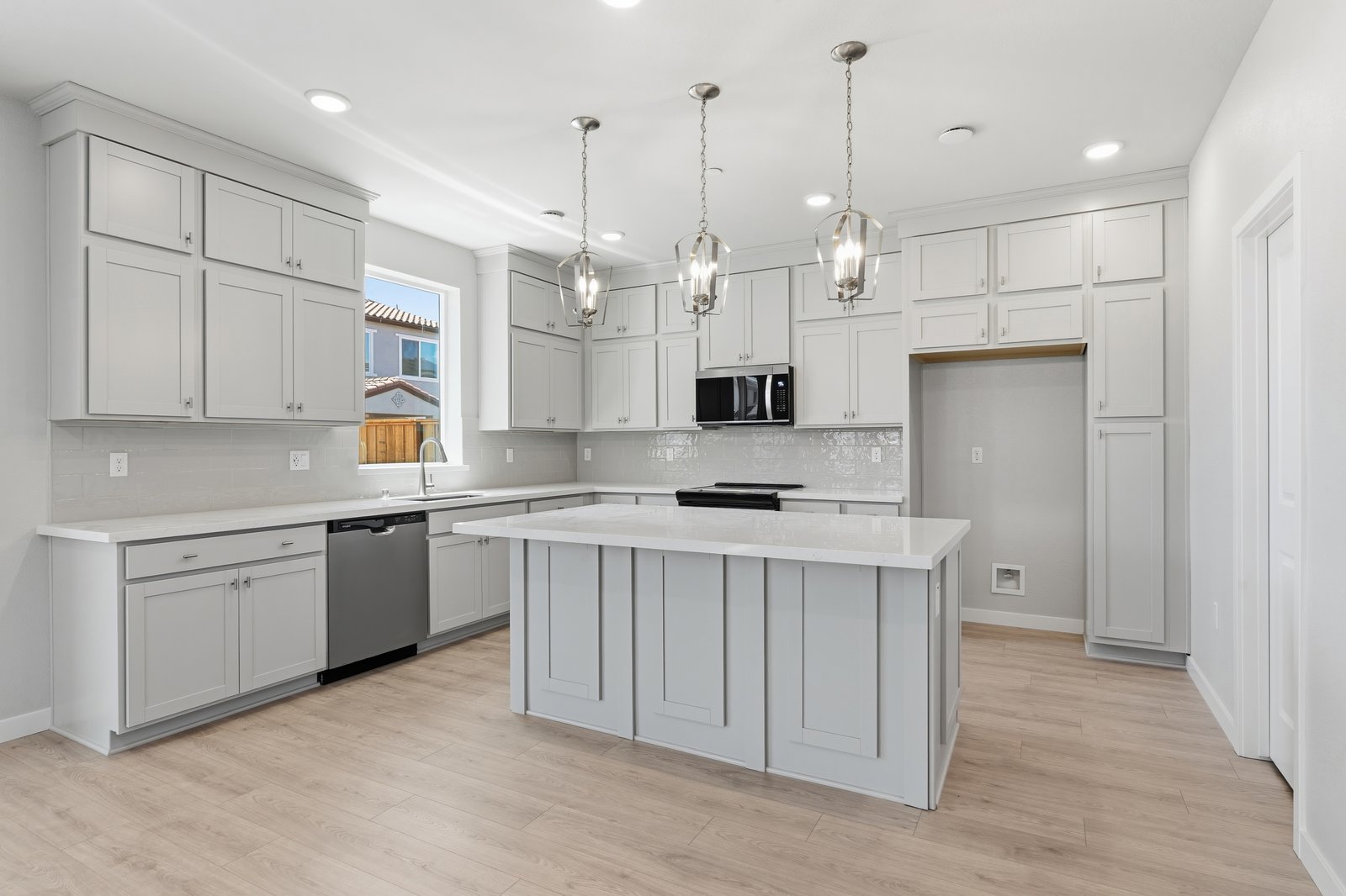kitchen with light gray cabinets, light wood floors, white counters, three brushed nickel pendants above island, and window