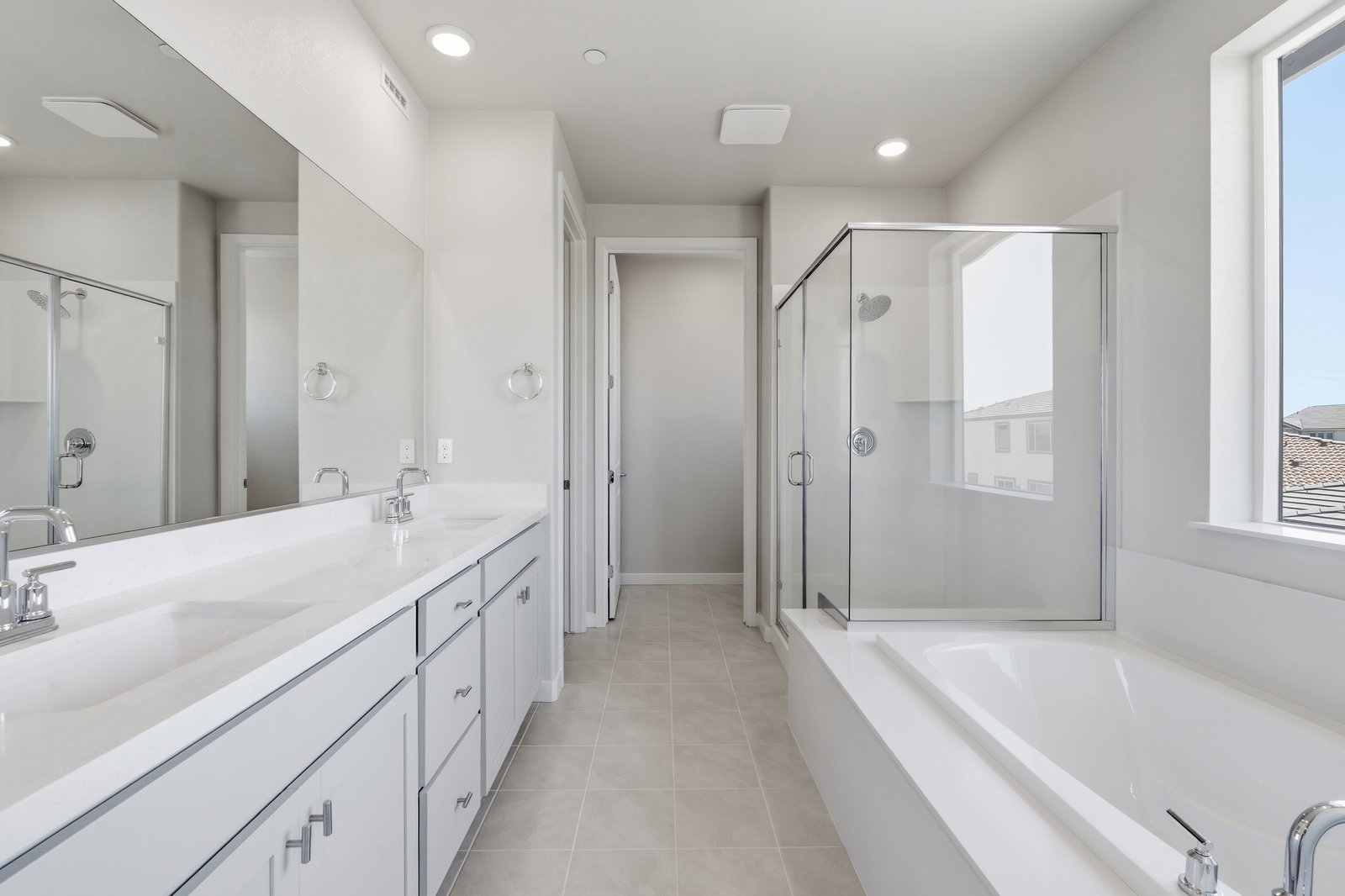 bathroom with light gray cabinets, white counters, two sinks, tan tile floor, walk-in shower, and window above tub