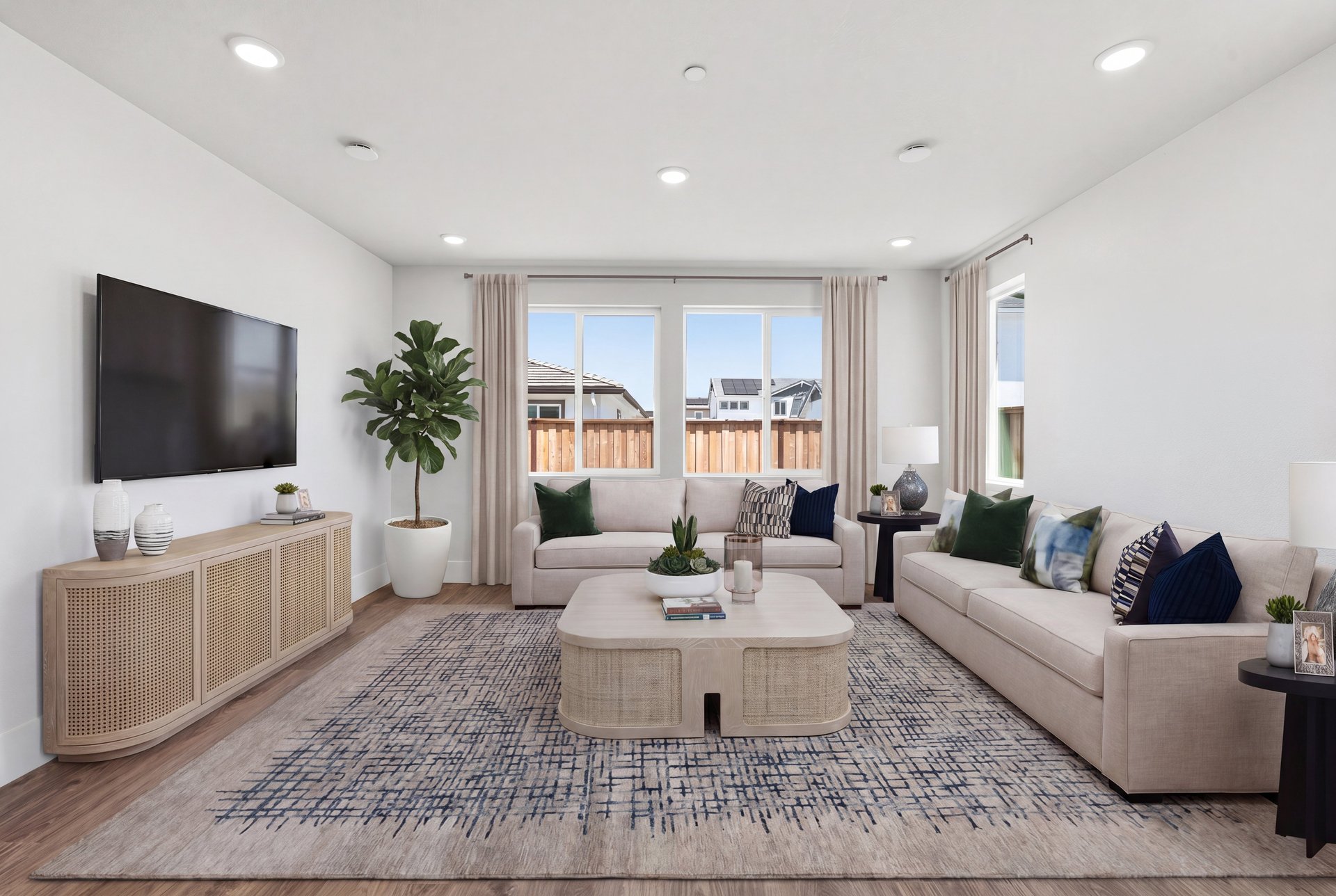 great room with wood floors, multiple windows, two tan sofas with green & blue pillows, and television on wall