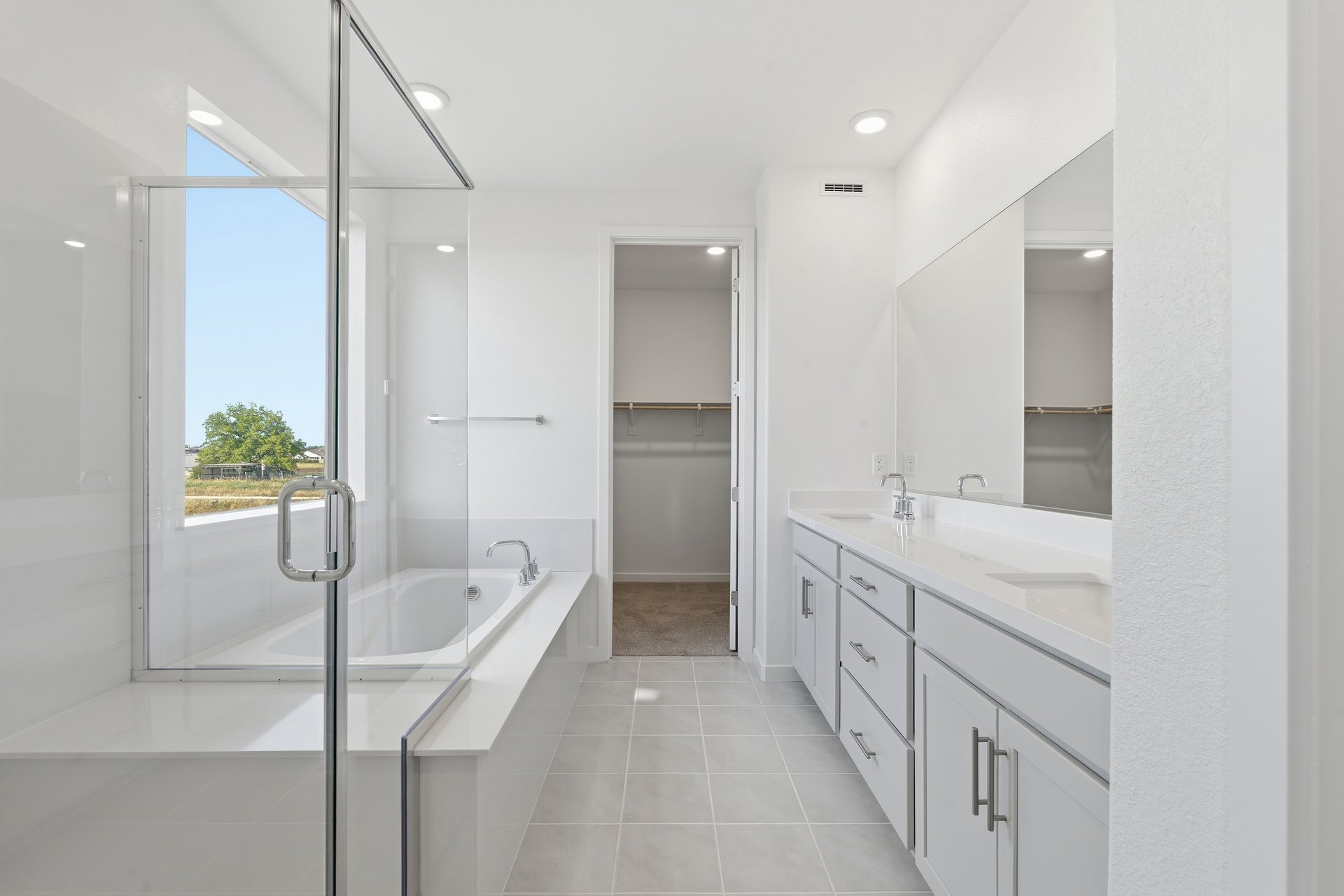 bathroom with white cabinets and counters, two sinks, walk-in shower, and large window above tub