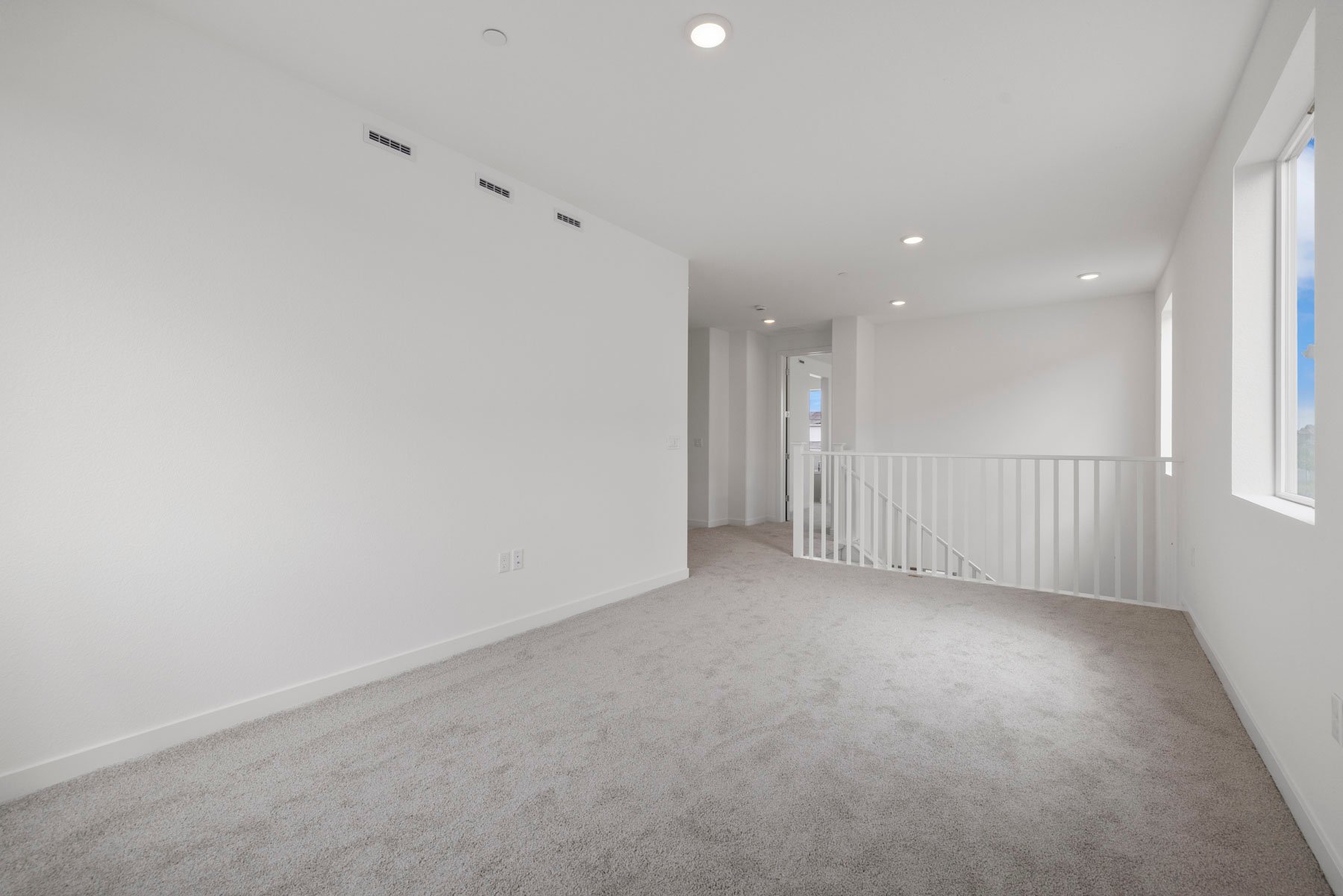 loft with tan carpet, white walls, windows, and view of stair rail leading down