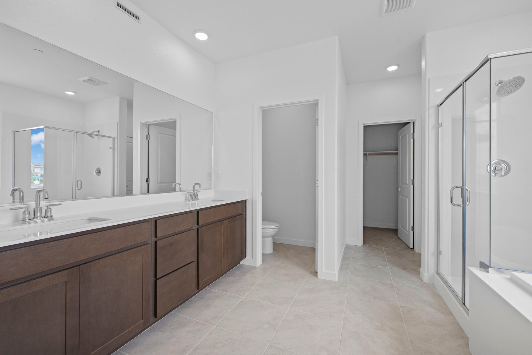 bathroom with brown cabinets, white counters, two sinks, and tan tile floors with walk-in shower