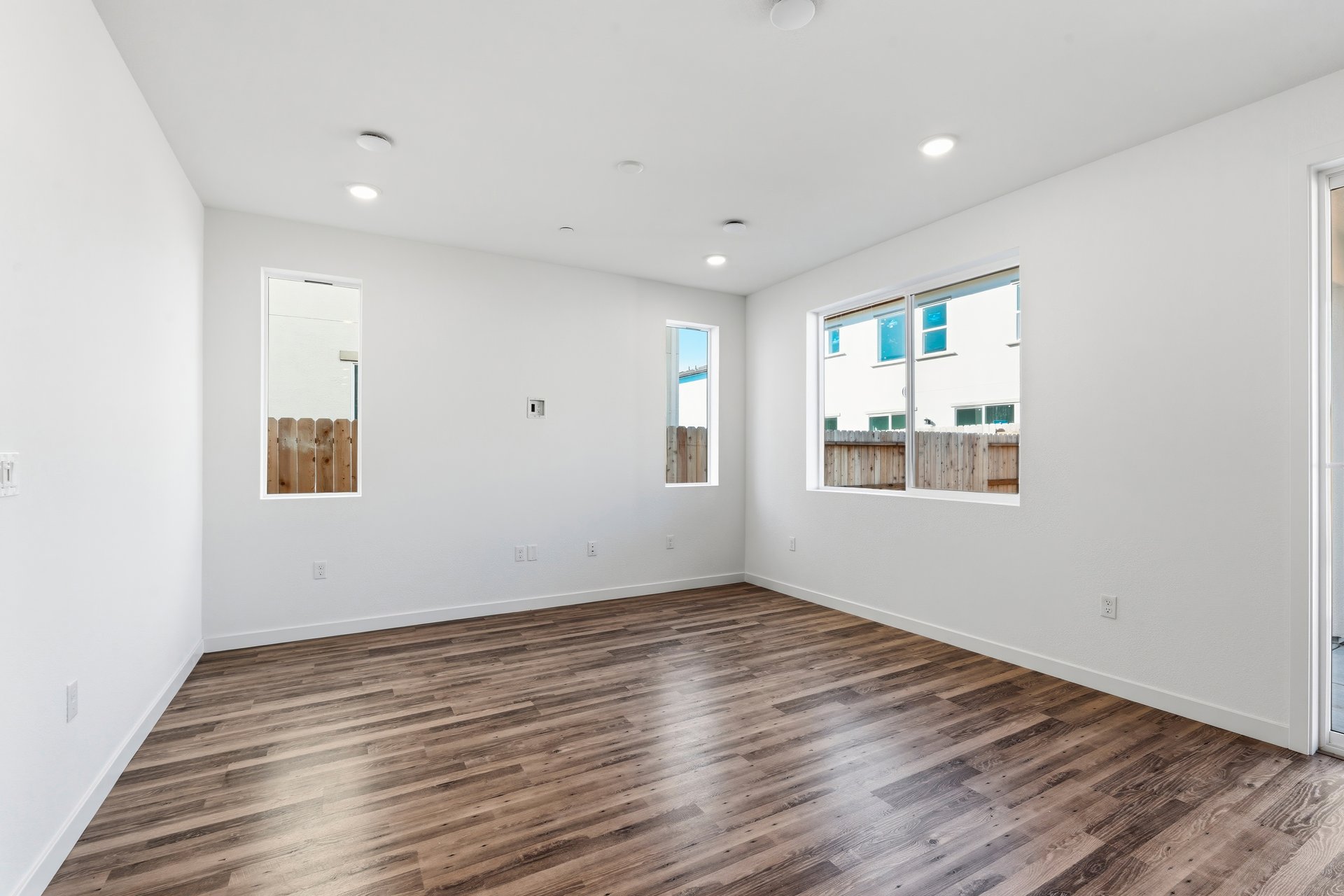 great room with wood floors, white walls, three windows looking out to the backyard, and recessed ceiling lights