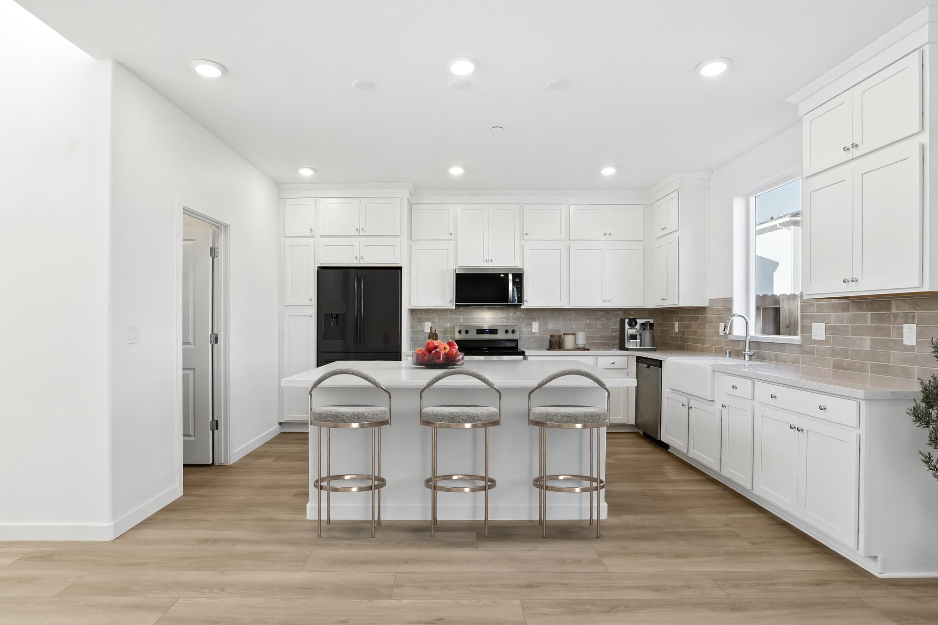 kitchen with white cabinets & counters, tan tile backsplash, wood floors, bowl of fruit on island, and three gray & gold barstools