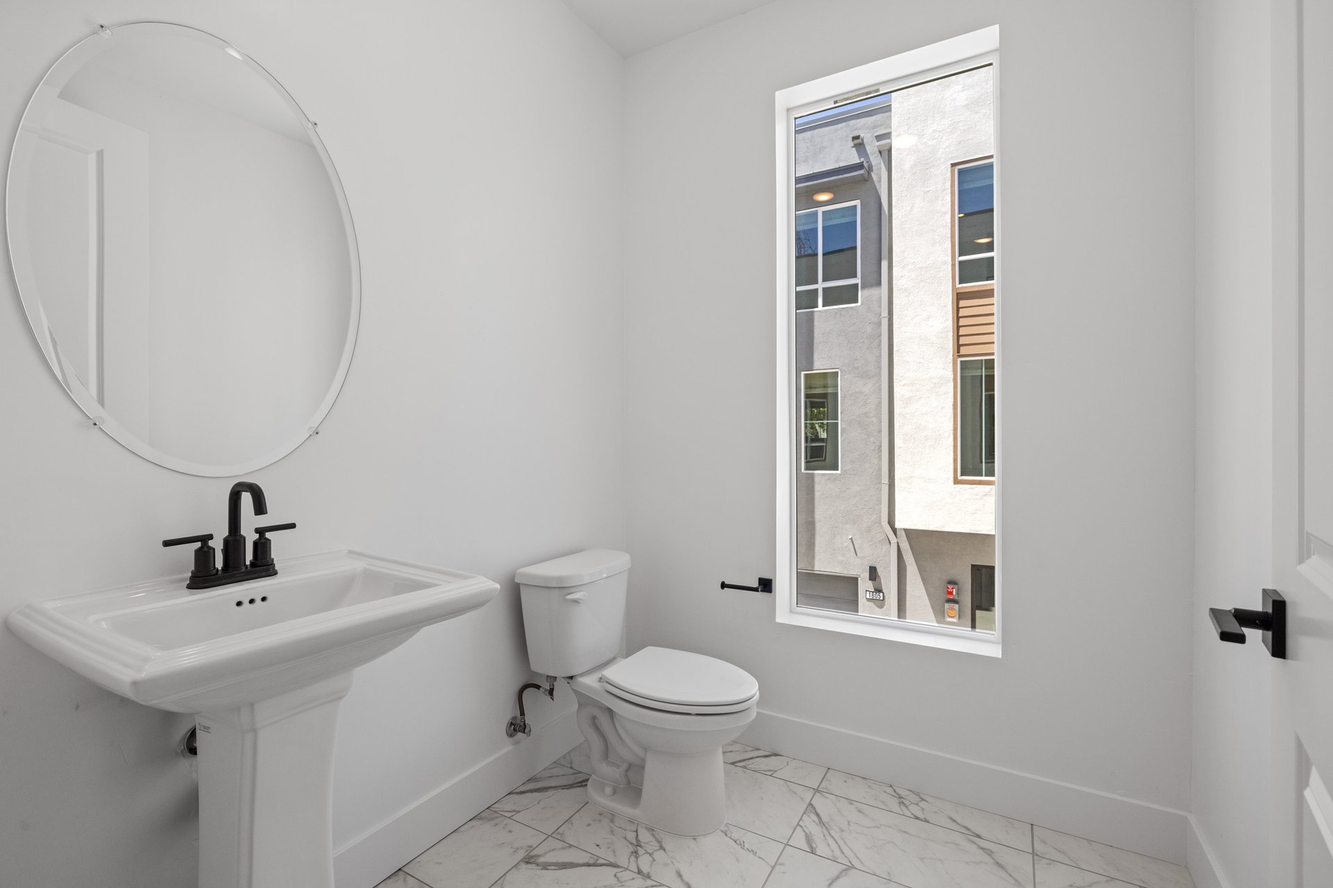 bathroom with window, round mirror above pedestal sink, and white & gray tile floor