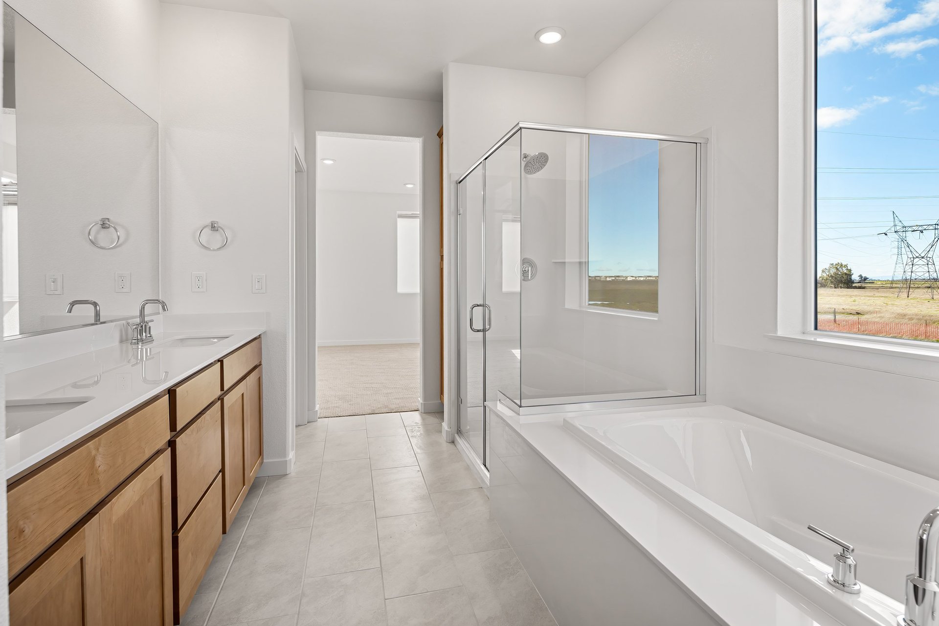 bathroom with wood cabinets, white counters, two sinks, light tan tile floors, window above large soaking tub, and walk-in shower