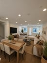great room with wood floors, white sectional sofa, wood dining table with six white chairs, and multiple windows