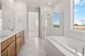 bathroom with wood cabinets, white counters, two sinks, light tan tile floors, window above large soaking tub, and walk-in shower