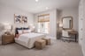 bedroom with tan carpet, window, mirror above wood desk, and white upholstered bed with two tan ottomans at foot